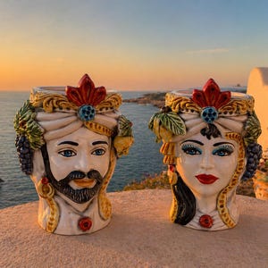 Puede incluir: Dos jarrones de cerámica con forma de cabeza, uno masculino y otro femenino, con detalles intrincados. El jarrón masculino tiene barba y bigote, mientras que el femenino tiene labios rojos y ojos azules. Ambos tienen tocados ornamentados.