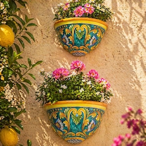 Op de afbeelding: Twee aan de muur gemonteerde keramische plantenbakken gevuld met roze en witte bloemen. De plantenbakken hebben een turquoise basis met gele en gouden sierlijke ontwerpen. De plantenbakken hangen aan een beige muur, met een citroenboom op de achtergrond.