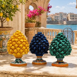 May include: Three ceramic pinecone sculptures in yellow, dark blue, and emerald green. Each piece features a glossy, textured surface with overlapping scales and sits on a decorative, hand-painted pedestal base with floral and geometric patterns.