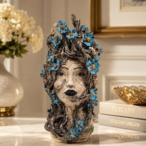 May include: A ceramic head sculpture featuring a weathered, cracked white finish with dark brown, branch-like hair textures. The sculpture is adorned with numerous small, vibrant blue flowers with yellow centers, and the face has dark lips.