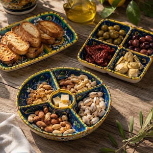 Può includere: Un set di piatti da portata in ceramica dipinti a mano con vivaci motivi floreali blu, gialli e verdi. Il set comprende un vassoio rettangolare con pane tostato, un piatto quadrato a quattro scomparti con olive e una ciotola rotonda a cinque sezioni riempita con noci e cubetti di formaggio.
