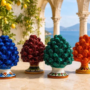 Op de afbeelding: Vier keramische dennenappelsculpturen in glanzend blauw, donkerrood, smaragdgroen en feloranje. Elk stuk heeft een textuur met gelaagde schubben en staat op een ronde voet die is versierd met handgeschilderde, kleurrijke bloemmotieven.