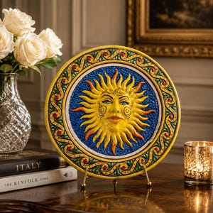 Puede incluir: Plato decorativo de cerámica con diseño de cara de sol. El plato tiene una cara amarilla, ojos azules y detalles en naranja. El centro es azul y el borde amarillo con detalles ornamentados. Se muestra en un soporte dorado.