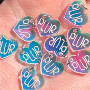 Könnte beinhalten: Eine Sammlung von irisierenden herzförmigen Anhängern. Jeder Anhänger hat eine kleine Schlaufe oben und das Wort "PLUR" in blauer Schrift. Die Anhänger werden in einer offenen Hand vor einem verschwommenen Hintergrund gehalten.