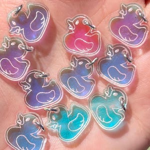 May include: A collection of small, iridescent duck-shaped charms in various colours, including pink, blue, and purple. Each charm has a white outline and is held in an open hand against a blurred green background.