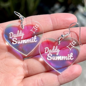 May include: A pair of iridescent heart-shaped earrings with the text "Daddy Summit" and a small star design.