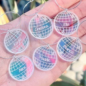 May include: A collection of personalized hoop earrings. Each earring features a globe design with a name, such as "Chelsea" or "Alex", in a clear, iridescent acrylic. The earrings have silver-colored hoops.