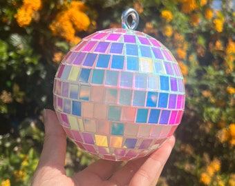 Può includere: Ornamento decorativo a forma di palla da discoteca con un design a mosaico. La palla ha una forma sferica con piastrelle iridescenti nei toni del rosa, blu e viola. Ha un anello di metallo argentato per appenderla. Lo sfondo presenta fiori gialli.