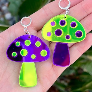 May include: A pair of mushroom-shaped earrings. One earring has a purple cap with green and purple circles, and a yellow-green stem. The other earring has a green cap with purple and green circles, and a purple stem. Both earrings have silver hoops.