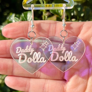 May include: A pair of iridescent heart-shaped earrings with the text "Daddy Dolla" on each earring.