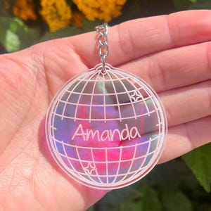 Op de afbeelding: Een ronde, heldere sleutelhanger met een wereldbolontwerp en de naam "Amanda" in witte letters. De sleutelhanger heeft een zilveren ketting en een kleurrijke, iriserende achtergrond. Het ontwerp staat tegen een achtergrond van gele bloemen.