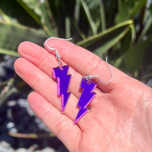 May include: A pair of purple lightning bolt earrings with silver hooks. The earrings are made of acrylic and have a mirrored finish.