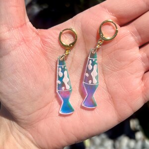 Op de afbeelding: Een paar goudkleurige oorringen met een lavalamp ontwerp. De oorbellen hebben een heldere, iriserende behuizing met witte, roze en blauwe accenten. De onderkant van de lavalamp is helderblauw, overgaand in paars. De oorbellen worden in een hand gehouden.