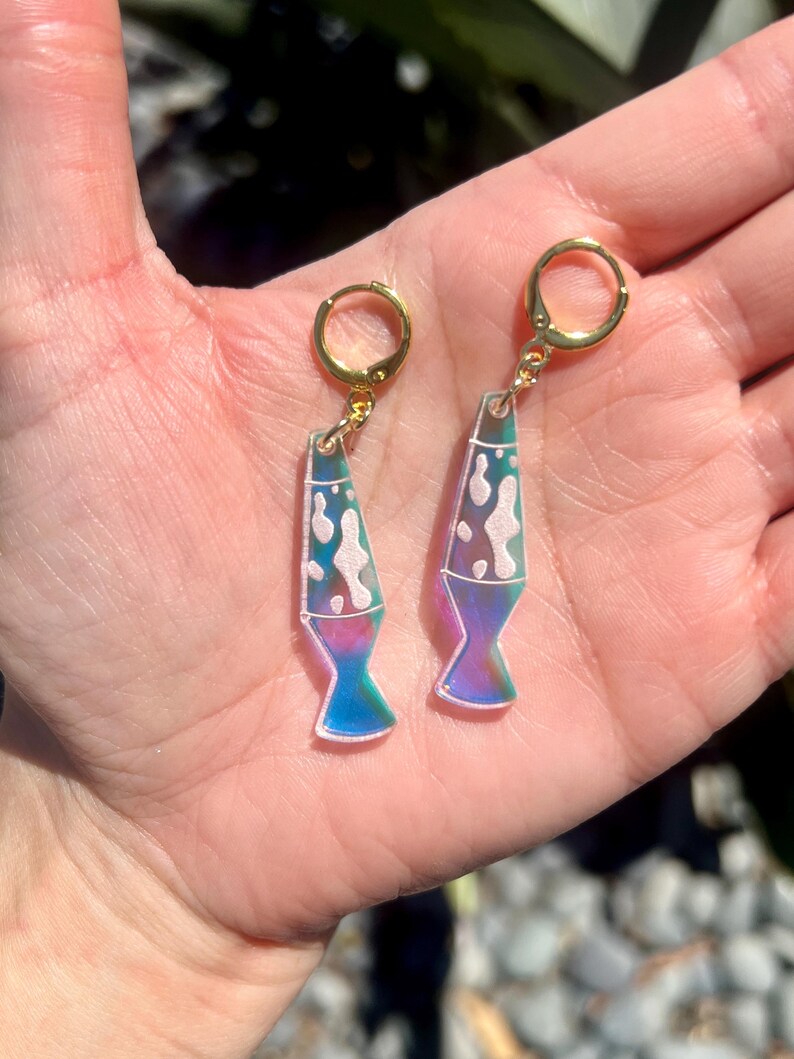 Op de afbeelding: Een paar goudkleurige oorringen met een lavalamp ontwerp. De oorbellen hebben een heldere, iriserende behuizing met witte, roze en blauwe accenten. De onderkant van de lavalamp is helderblauw, overgaand in paars. De oorbellen worden in een hand gehouden.