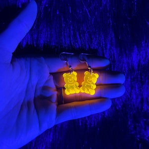 May include: A pair of yellow gummy bear shaped earrings. The earrings are glowing under a black light.