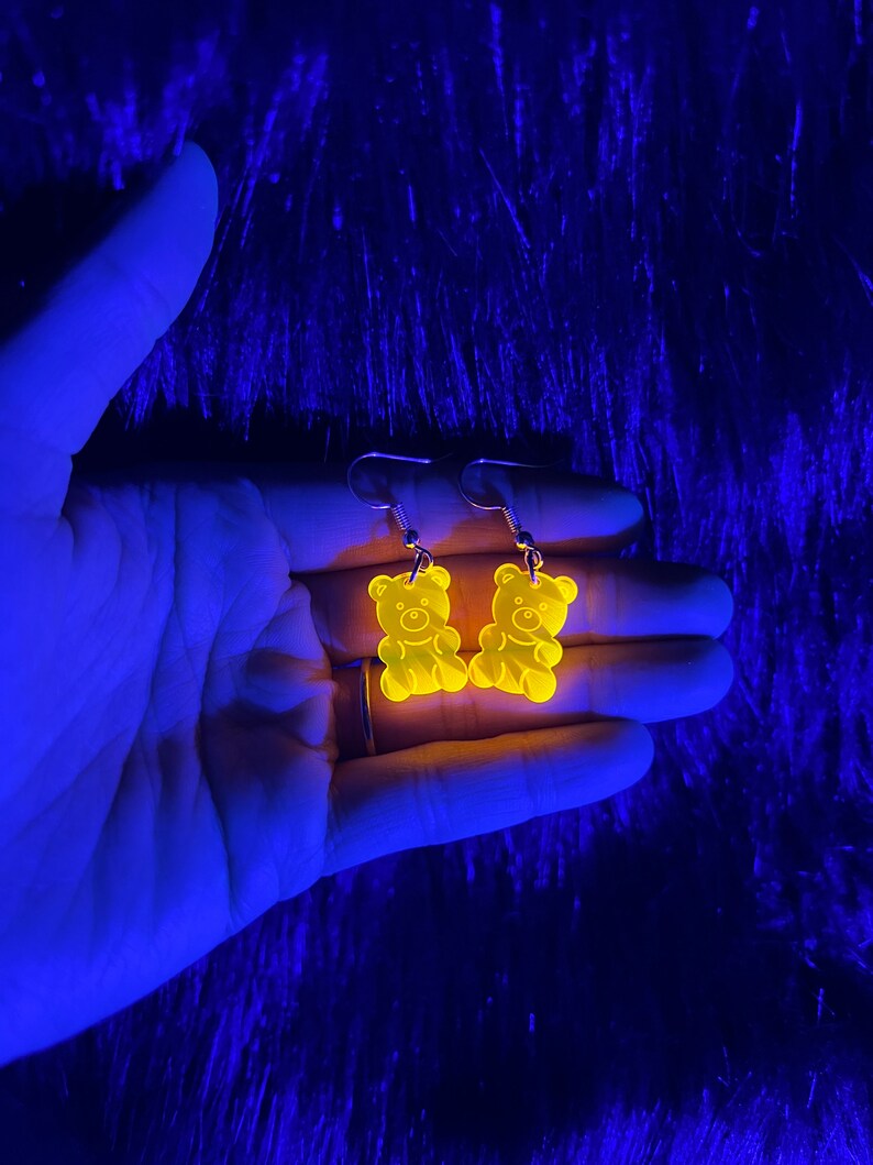 May include: A pair of yellow gummy bear shaped earrings. The earrings are glowing under a black light.