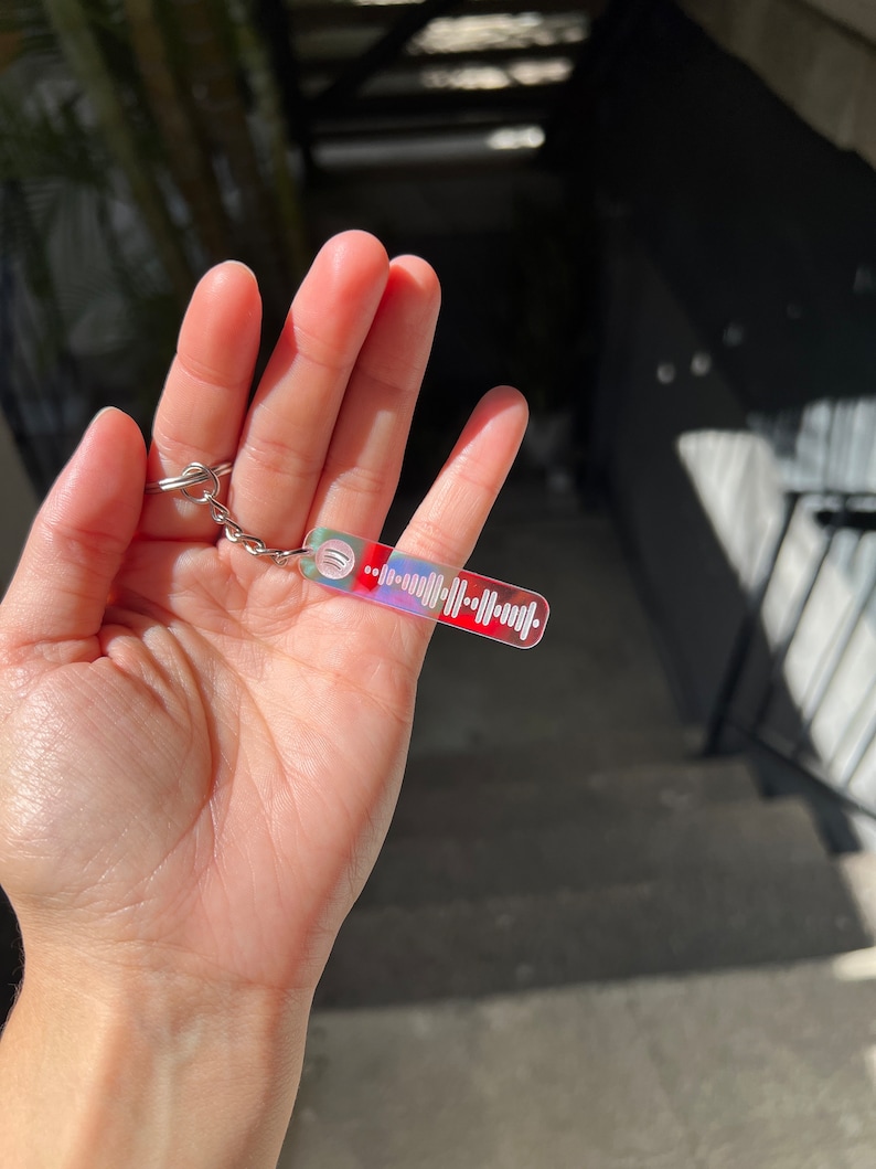 Op de afbeelding: Een kleurrijke, iriserende sleutelhanger met een Spotify-code erop gedrukt. De sleutelhanger is gemaakt van helder acryl en heeft een zilveren metalen ring.