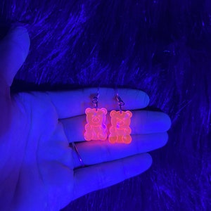 May include: A pair of orange gummy bear shaped earrings. The earrings are made of clear acrylic and glow under a black light.