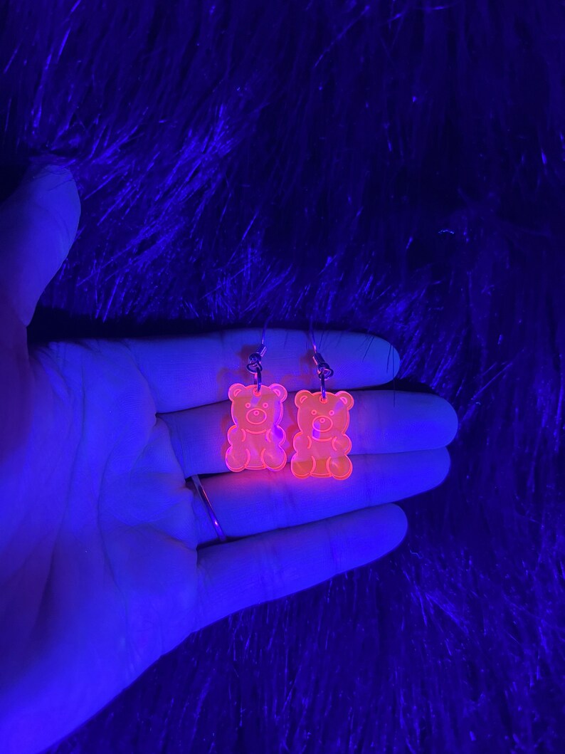 May include: A pair of orange gummy bear shaped earrings. The earrings are made of clear acrylic and glow under a black light.