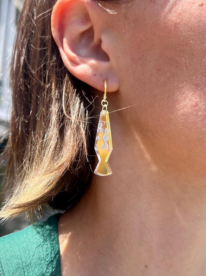 Op de afbeelding: Goudkleurige lava lamp oorbellen met een geel en wit ontwerp. De oorbellen hangen aan een gouden oorring. Een leuk en uniek accessoire.
