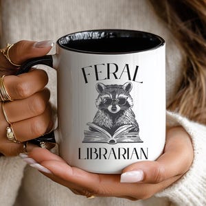May include: White ceramic mug with a black interior and handle. The mug features the text "FERAL LIBRARIAN" with an illustration of a raccoon wearing glasses and reading a book. The mug is held by a person.