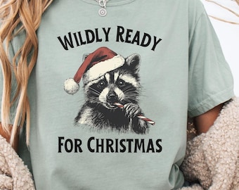 Può includere: T-shirt verde chiaro con la scritta "WILDLY READY FOR CHRISTMAS" sopra e sotto un'illustrazione dettagliata di un procione con un cappello da Babbo Natale e un bastoncino di zucchero. Il procione è bianco e nero.