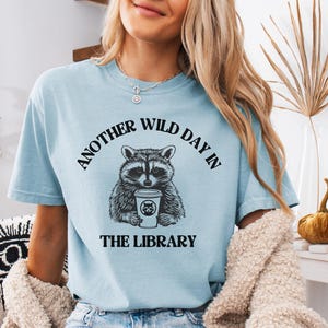 Op de afbeelding: Lichtblauw T-shirt met de tekst "ANOTHER WILD DAY IN THE LIBRARY" boven en onder een wasbeer-afbeelding die een koffiekopje vasthoudt. De wasbeer-afbeelding is zwart-wit. Het shirt is gemaakt van een zacht materiaal.