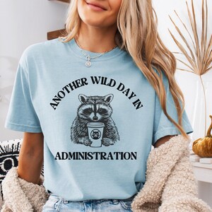 以下が含まれることがあります： 「ANOTHER WILD DAY IN ADMINISTRATION」の文字が、コーヒーカップを持ったアライグマのグラフィックの上と下に配置された、水色のTシャツ。アライグマは白黒で、テキストは黒色です。このシャツは柔らかい素材で作られています。