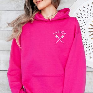 Puede incluir: Una sudadera con capucha de color rosa brillante con un gran bolsillo delantero. La sudadera tiene un cordón blanco y la palabra "TAYLOR" está bordada en la parte delantera con dos remos cruzados.