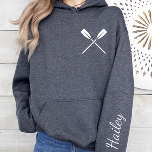 Puede incluir: Sudadera con capucha gris oscuro con un diseño de remos blancos cruzados en el pecho y el nombre "Hailey" impreso en la manga. La sudadera tiene un bolsillo delantero y una capucha.