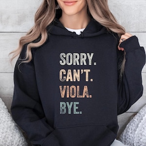 以下が含まれることがあります： 「SORRY. CAN'T. VIOLA. BYE.」のテキストがヴィンテージ風にプリントされた黒いパーカー。テキストはベージュ、オレンジ、グリーンで、フードにはドローコード、前面にはポケットが付いています。