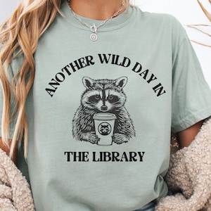 May include: Sage green t-shirt featuring a raccoon holding a coffee cup. The shirt has the text "ANOTHER WILD DAY IN THE LIBRARY" in a black, arched font above and below the raccoon graphic. The raccoon graphic is black and white.