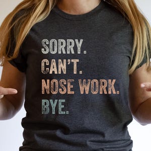 May include: A dark heather grey t-shirt with the text "SORRY. CAN'T. NOSE WORK. BYE." in a vintage style font. The text is in shades of pink, orange, and blue.