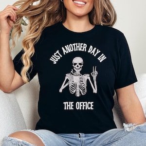Op de afbeelding: Zwart T-shirt met een skelet dat een kopje koffie vasthoudt en een peace-teken maakt. De tekst op het shirt luidt "Just another day in the office."