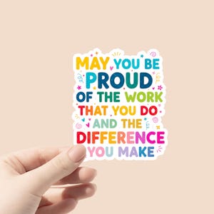 May include: A colorful sticker with the text "MAY YOU BE PROUD OF THE WORK THAT YOU DO AND THE DIFFERENCE YOU MAKE." The sticker has a white border and is held by a hand against a light peach background.