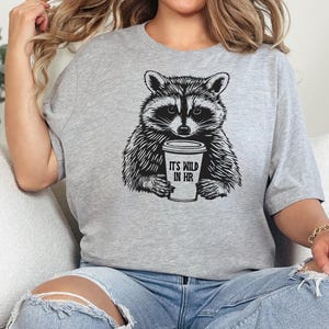 May include: A light grey t-shirt with a black and white drawing of a raccoon holding a coffee cup. The cup has the text "IT'S WILD IN HR". The t-shirt is a crew neck style.