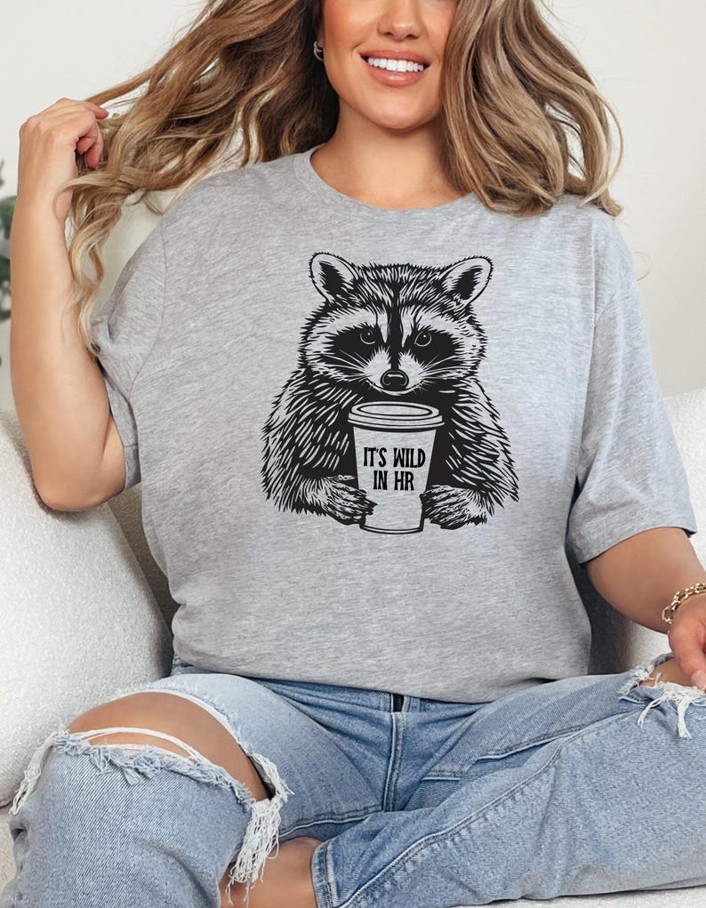 May include: A light grey t-shirt with a black and white drawing of a raccoon holding a coffee cup. The cup has the text "IT'S WILD IN HR". The t-shirt is a crew neck style.