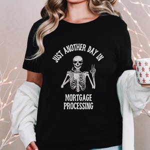 May include: Black t-shirt with a white skeleton holding a coffee cup and making a peace sign. The text on the shirt reads "Just another day in mortgage processing."