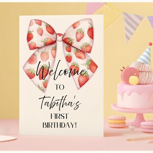以下が含まれることがあります： イチゴのリボンのデザインと「Welcome to Tabitha's First Birthday！」の文字が入った誕生日パーティーの招待状。パーティーハットとマカロンが付いたピンク色のケーキも表示されています。