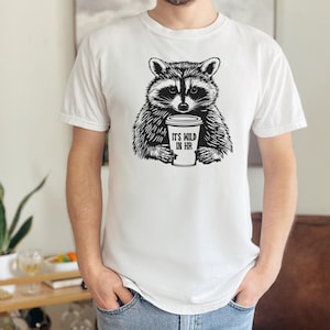 May include: White t-shirt with a black and white raccoon graphic holding a coffee cup. The cup reads "IT'S WILD IN HR". The raccoon design is a central feature of the shirt.