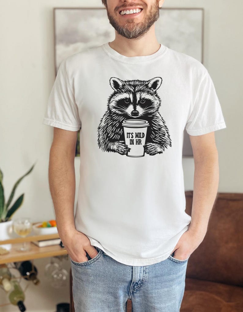 May include: White t-shirt with a black and white raccoon graphic holding a coffee cup. The cup reads "IT'S WILD IN HR". The raccoon design is a central feature of the shirt.