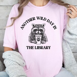 Peut inclure: T-shirt rose clair avec une illustration en noir et blanc d'un raton laveur tenant une tasse de caf&eacute;. Le texte au-dessus du raton laveur se lit "ANOTHER WILD DAY IN" et en dessous "THE LIBRARY".