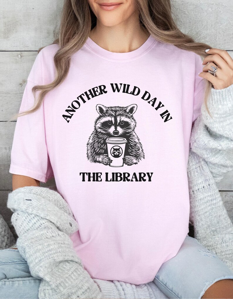 Peut inclure: T-shirt rose clair avec une illustration en noir et blanc d'un raton laveur tenant une tasse de caf&eacute;. Le texte au-dessus du raton laveur se lit "ANOTHER WILD DAY IN" et en dessous "THE LIBRARY".