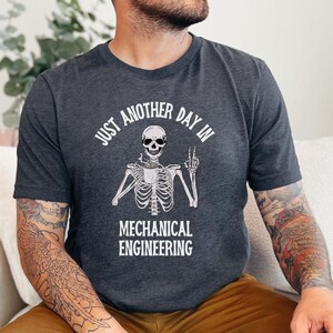 May include: A dark gray t-shirt with a white skeleton holding a coffee cup and the text "Just another day in mechanical engineering".