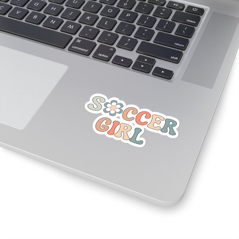 May include: A sticker with the words "Soccer Girl" in a retro font with a flower design. The sticker is in shades of pink, blue, and green.
