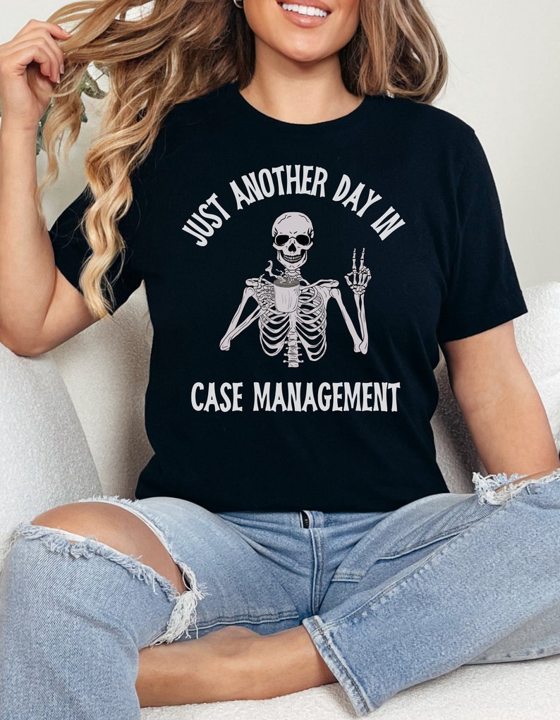 May include: Black t-shirt with a white graphic of a skeleton holding a coffee cup and giving a peace sign. The text "Just another day in case management" is printed on the shirt.