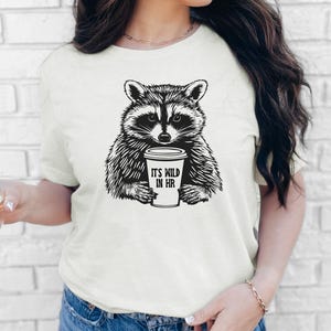 May include: A cream-coloured t-shirt with a black and white raccoon illustration holding a coffee cup. The cup reads "IT'S WILD IN HR". The shirt is a short-sleeved crew neck.