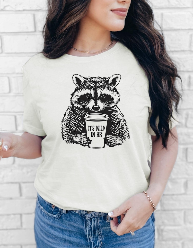 May include: A cream-coloured t-shirt with a black and white raccoon illustration holding a coffee cup. The cup reads "IT'S WILD IN HR". The shirt is a short-sleeved crew neck.