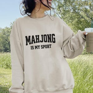 May include: A beige sweatshirt with the text "MAHJONG IS MY SPORT" in black block letters. The sweatshirt is a casual style, suitable for everyday wear. The image is taken outdoors with a green background.
