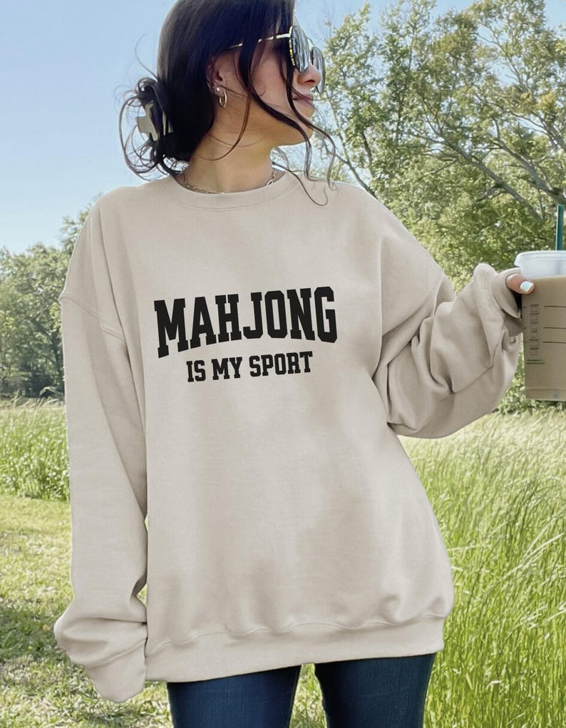 May include: A beige sweatshirt with the text "MAHJONG IS MY SPORT" in black block letters. The sweatshirt is a casual style, suitable for everyday wear. The image is taken outdoors with a green background.